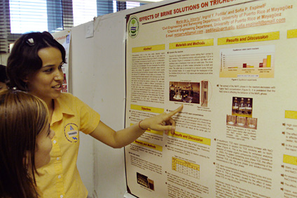 La estudiante graduada María de Lourdes Irizarry muestra a una companera el afiche de su investigación.