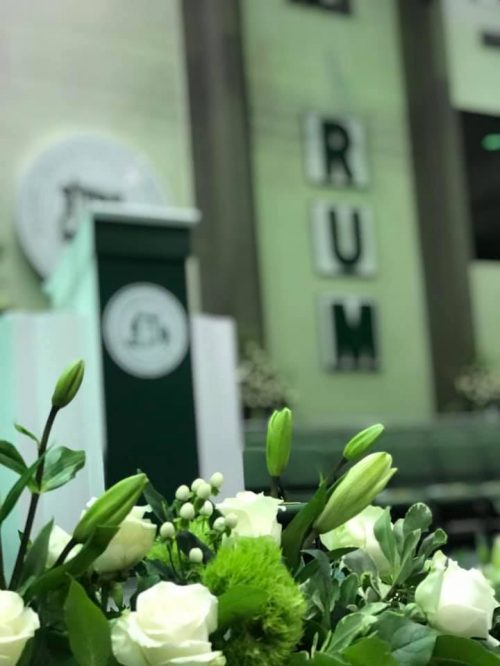 Imagen mostrando flores en primer plano y en segundo plano el podio con el sello del recinto y las siglas RUM