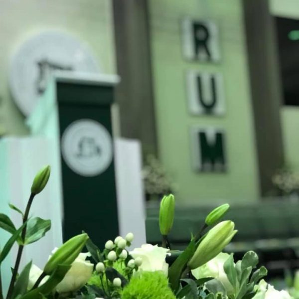 Imagen mostrando flores en primer plano y en segundo plano el podio con el sello del recinto y las siglas RUM