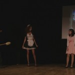 Cast members for the student presented play of The Rocky Horror Picture show on stage with the film being displayed in the background. There is a couple standing together, a maid and a man holding a duster.