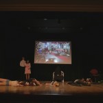 Cast members for the student presented play of The Rocky Horror Picture show on stage. A couple is standing to the left of the stage while everyone else is collapsed on the floor of the stage.