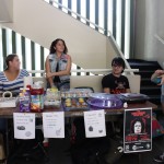 The table of the EDSA Book & Bake Sale is lined up with promo posters for a theater event, as well as the prices for the items being sold. The EDSA team members are interacting with each other.