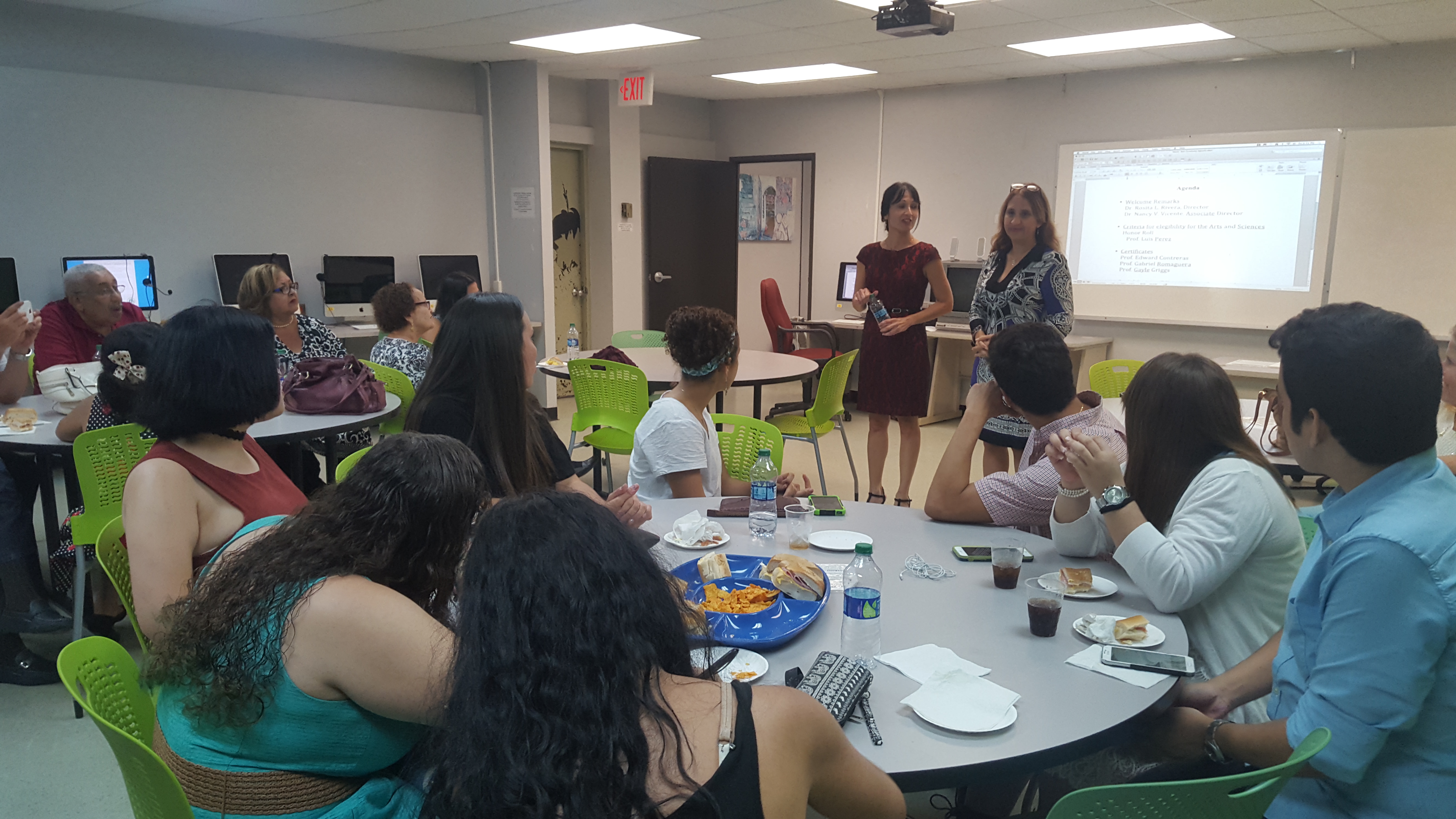 Two college professors speaking in front of a room at the Honor Roll Students Celebration. Students are sitting at the tables and there are snack trays on top of the tables.