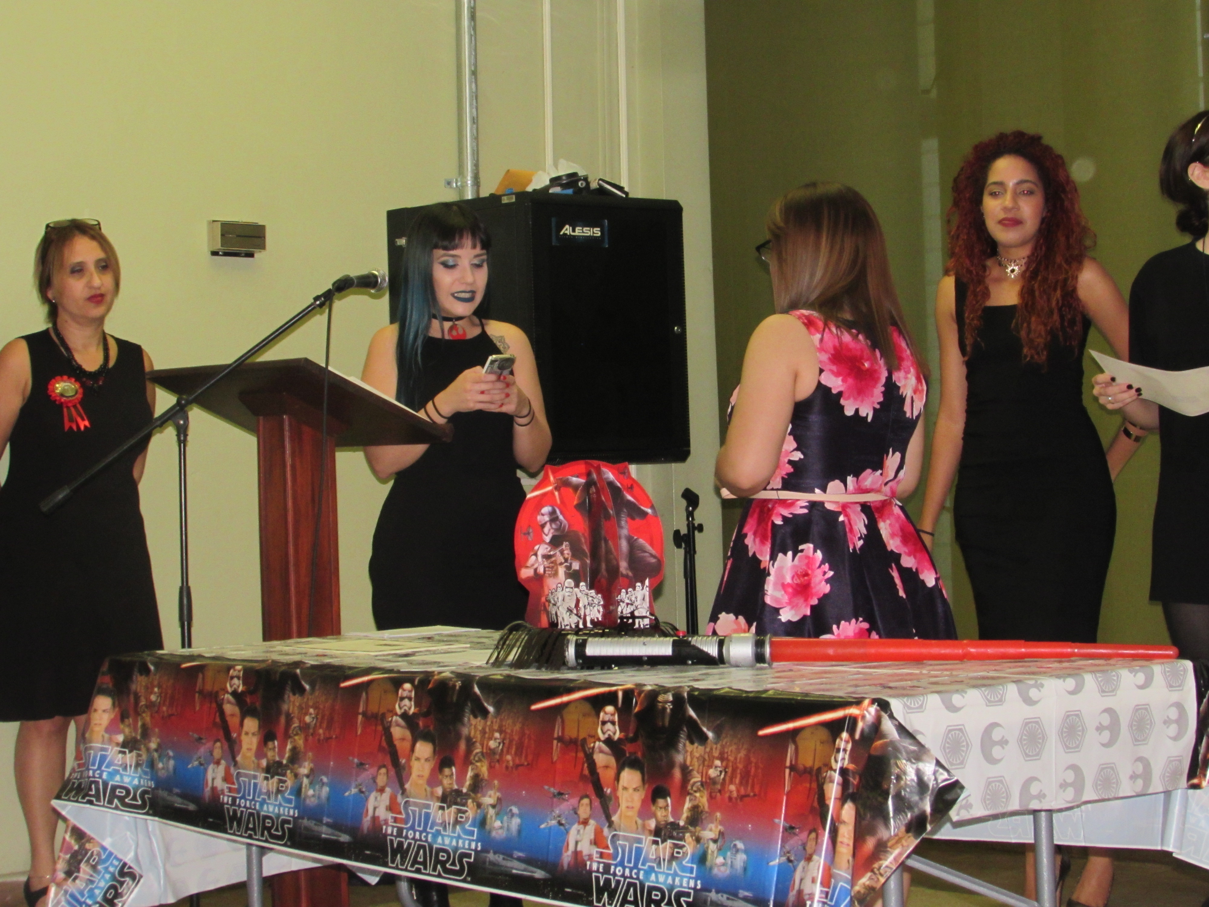 Former President Ivette López passing the baton to Ana Carrero as the new PCSA President. The president is by the podium reading from a smartphone and a table is lined up with Star Wars decoration.