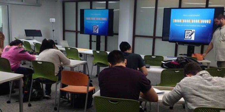edcelgric Several students are sitting by tables while working diligently on their papers.
