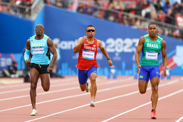 La imagen muestra a tres atletas corriendo en la pista y en el centro se destaca Diego Andrés González Salgado, estudiante del Recinto Universitario de Mayagüez en los Juegos Panamericanos 2023.