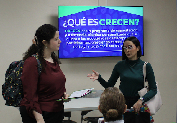 Portada CRECEN Dos participantes en la Feria de empleo de CRECEN en el RUM conversan. Detrás de ellas, una pantalla electrónica con información del programa.