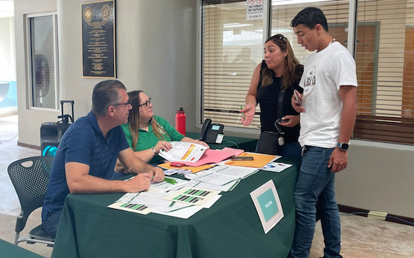 Dos representantes colegiales sentados en una mesa conversan con un estudiante y su madre, de pie, en la Feria de Orientaciones.