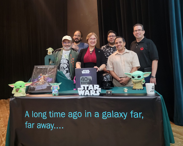 Grupo de organizadores y conferenciantes de la semana dedicada a las películas Star Wars en el RUM, de pie frente a una mesa con artículos y un cartel alusivos a la saga.