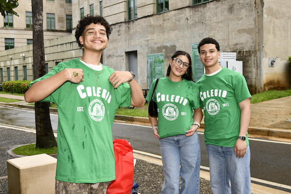 Tres estudiantes de nuevo ingreso posan para la cámara con sus camisetas verdes que dicen Colegio junto al emblema institucional.