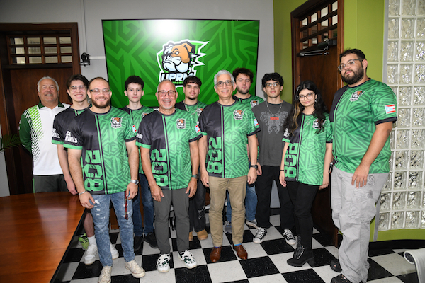 Varios integrantes de la organización estudiantil de videojuegos UPRM Esports, junto al rector del RUM, doctor Agustín Rullán Toro, el profesor José ‘Tito’ Estévez, y el entrenador Félix ‘Pompi’ Vega. Todos de pie, con sus uniformes y con el logo del colectivo proyectado al fondo en una pantalla.