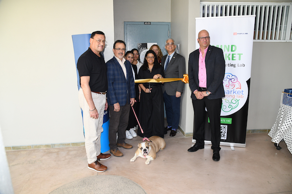 Personal docente y administrativo del Recinto Universitario de Mayagüez inaugura las instalaciones del NeuroLab MindMarket Lab en el Edificio de Empresas. Todos de pie frente a la puerta del laboratorio en el corte de cinta, con la mascota Jane.