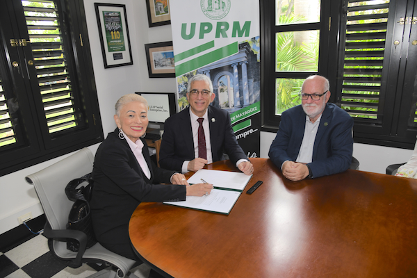 El Colegio de Ciencias Agrícolas del Recinto Universitario de Mayagüez firmó un acuerdo de colaboración con la escuela LEAP STEAM+ Agriculture Sciences. De izquierda a derecha: los doctores Gloria Bonilla, principal oficial ejecutiva de LEAP Social Enterprise; Agustín Rullán Toro, rector del RUM; y Raúl Macchiavelli, decano y director del CCA. Los tres sentados en una mesa en la Oficina del Rector con los documentos para firmar. En la parte de atrás, un cartel con el logo del Recinto.