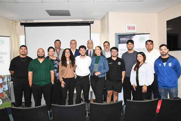 Foto grupal de los participantes del nuevo ciclo del Programa Ejecutivo de Gestión del Deporte que se desarrolla mediante la colaboración entre la Federación Puertorriqueña de Fútbol, la Federación Internacional de Fútbol y el Centro Internacional de Estudios del Deporte (CIES) en el Recinto Universitario de Mayagüez. Todos de pie y a los lados carteles con los logos de las entidades.