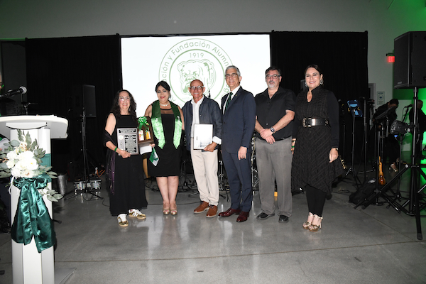 Como parte de los actos, se rindió homenaje a dos colegiales distinguidos: el agrónomo Douglas Candelario Nazario y la doctora Luisa Rosario Seijo Maldonado, al centro, acompañados por el rector, varios decanos y la presidenta de la Asociación y Fundación Alumni Colegial. Al fondo una pantalla proyecta el logo de la Asociación en colores blanco y verde.