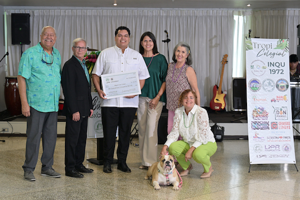 Personal de la Oficina de Exalumnos y el rector del RUM en la entrega de la beca uno de los estudiantes, al centro sosteniendo el cheque simbólico. Todos de pie, una de las administradoras en cuclillas junto a la mascota Jane.