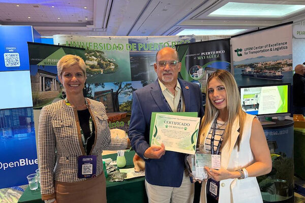 La doctora Ingrid Y. Padilla Cestero, primera desde la izquierda, acompañada por dos personas del grupo que representó al Recinto Universitario de Mayagüez en la más reciente Convención de Industriales de Puerto Rico 2026. Los tres de pie frente a la mesa informativa de la institución.