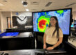 Katiria Colón Rivera, estudiante del Recinto Universitario de Mayagüez, obtuvo la prestigiosa beca Ernest F. Hollings que otorga la Administración Nacional Oceánica y Atmosférica y representa al RUM este verano en un internado en el Centro Nacional de Huracanes en Miami. De pie frente a un monitor grande de computadora que proyecta una gráfica de un huracán en las oficinas del Centro.
