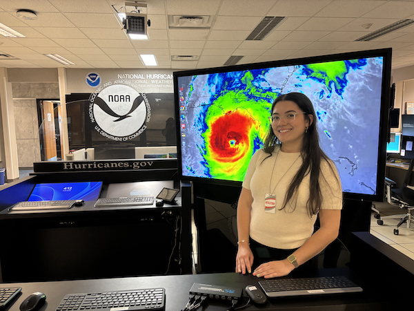 Katiria Colón Rivera, estudiante del Recinto Universitario de Mayagüez, obtuvo la prestigiosa beca Ernest F. Hollings que otorga la Administración Nacional Oceánica y Atmosférica y representa al RUM este verano en un internado en el Centro Nacional de Huracanes en Miami. De pie frente a un monitor grande de computadora que proyecta una gráfica de un huracán en las oficinas del Centro.