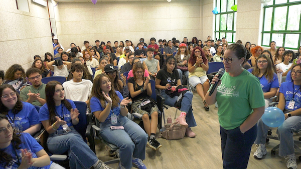 Una exalumna colegial se dirige a un grupo de recién admitidos al Recinto Universitario de Mayagüez (RUM), de pie, al centro y los estudiantes sentados la observan.
