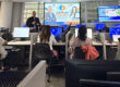 Al fondo, Jesús “Nesty” Delgado, de TEAM 1144, ofreció un taller de Inteligencia Artificial al personal del Colegio de Administración de Empresas del RUM. El conferenciante está de pie al lado de una pantalla con su presentación y lo observan varios participantes sentados en un salón con computadoras.