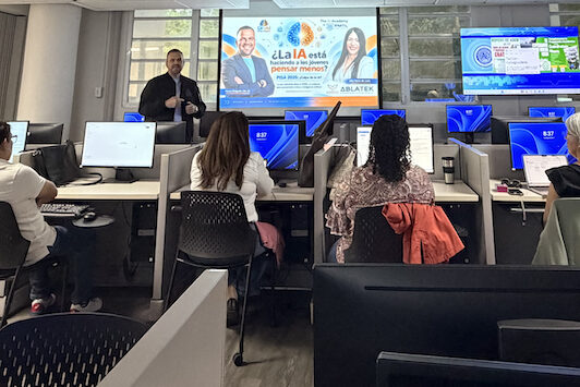 Portada Al fondo, Jesús “Nesty” Delgado, de TEAM 1144, ofreció un taller de Inteligencia Artificial al personal del Colegio de Administración de Empresas del RUM. El conferenciante está de pie al lado de una pantalla con su presentación y lo observan varios participantes sentados en un salón con computadoras.